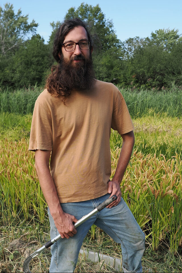 Orion Blake holding farm implements and wearing a brown shirt as he stands in a rice field on his farm